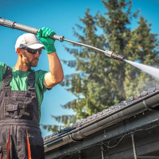 Technician clearing packed leaves from second-story gutters beside a tree-lined suburban home